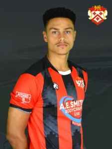 Portrait of a male footballer wearing a red and black striped jersey with a team crest on the chest, posed against a dark background. Crest appears in the upper right corner.