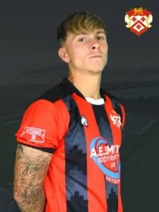 Portrait of a young man in a red and black striped football jersey, with team crest, tattoos on his forearm, against a dark background.