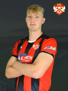 Portrait of a young man in a red and black striped football jersey with arms folded, team crest on the chest.