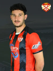 Portrait of a young man in a red and black striped football jersey with team crest on chest, horizon background and dim cityscape.