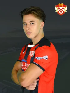 Portrait of a young male footballer with arms crossed, wearing a red and black jersey with a crest on the chest against a dark background.
