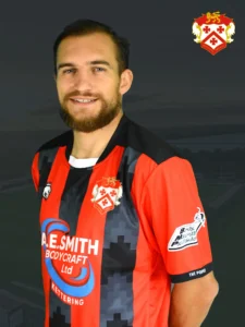 Portrait of a smiling man wearing a red and black striped football jersey with club crest in the top right, dark background behind him