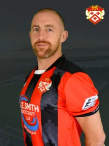 Portrait of a man in a red and black striped sports jersey with a team crest on the chest against a dark background.