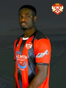 Portrait of a football player wearing a red and black Kettering Town FC jersey, with the club crest in the upper right and a dark backdrop behind him.