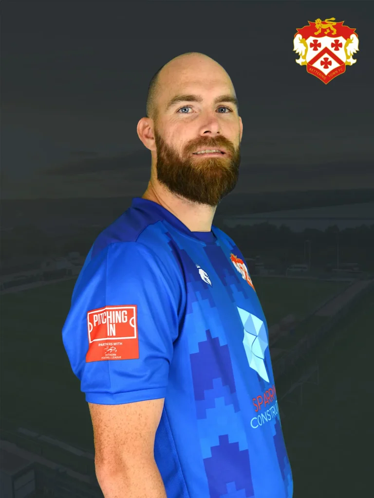 Portrait of a bearded man in a blue sports jersey with a red sleeve patch, set against a dark stadium backdrop.