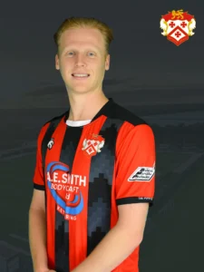 Smiling man in a red-and-black striped soccer jersey with a club crest on the chest, against a dark background.