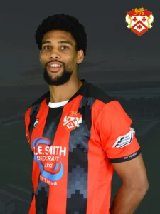 Smiling man wearing a red and black striped football jersey with a crest on the chest, standing against a dark background. Crest visible in the top-right corner.