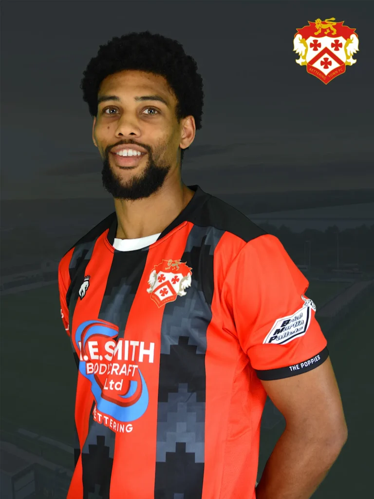 Smiling man wearing a red and black striped football jersey with a crest on the chest, standing against a dark background. Crest visible in the top-right corner.