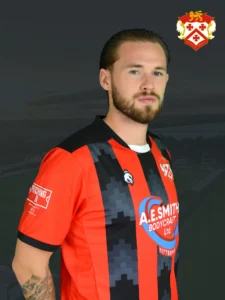 Portrait of a man with short hair and beard wearing a red and black striped soccer jersey, with a team crest visible on the chest and a logo on the front, standing outdoors at dusk with a dark sky background.