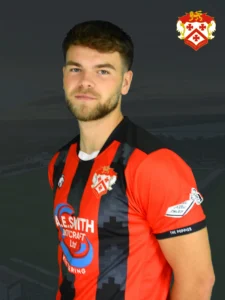 Portrait of a man in a red and black striped soccer jersey with a club crest on the chest, against a dark backdrop.