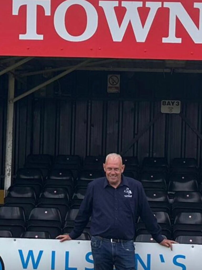 KTFC_BM_Pallets | Kettering Town FC Smiling man in a navy shirt and jeans stands in front of empty stadium seats under a red sign, with a no-smoking sign and'Bay 3' visible.