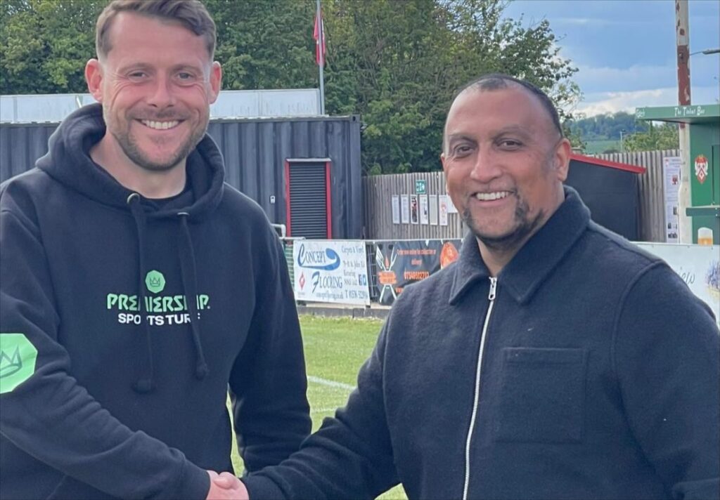 premiership_sports_turf_Image | Kettering Town FC Two men smiling and shaking hands on a grassy sports field, wearing dark jackets, with banners and fences in the background.