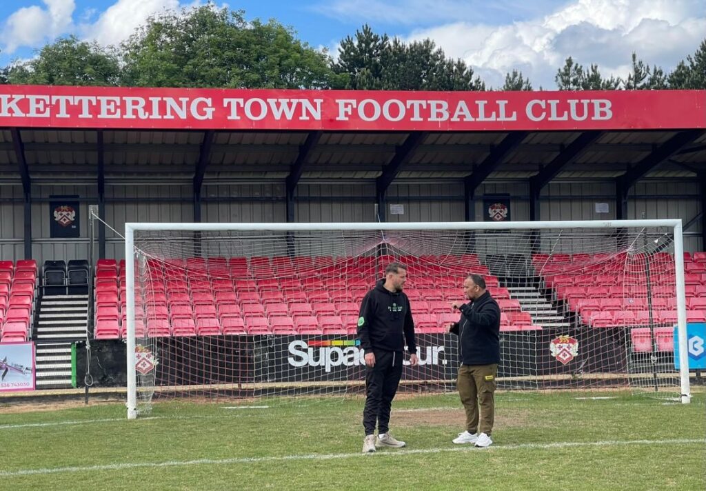 premiership_sports_turf_KTFC_Stadium | Kettering Town FC Two men standing and talking in front of a football goal on a grassy pitch at Kettering Town FC stadium with red seats behind them.