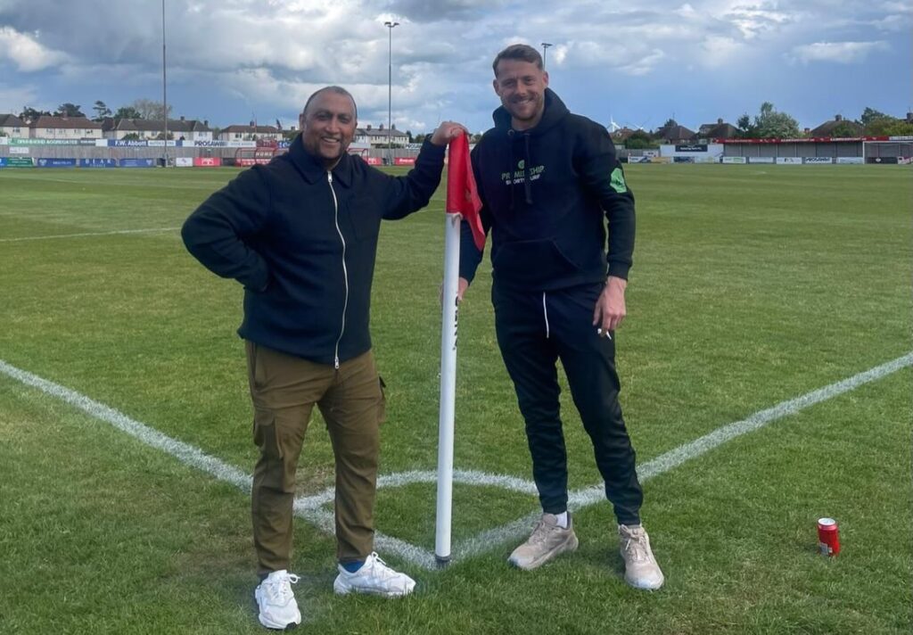 premiership_sports_turf_sponsorship | Kettering Town FC Two men pose by a red corner flag on a grassy soccer field, smiling at the camera with the flag between them.
