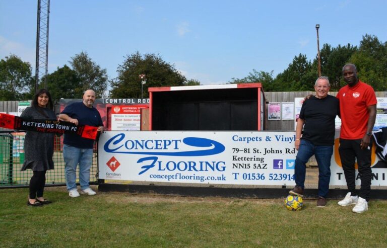 Five people stand along a fence with sponsor banners, including Concept Flooring, at a football ground; a colorful soccer ball rests on the grass nearby.