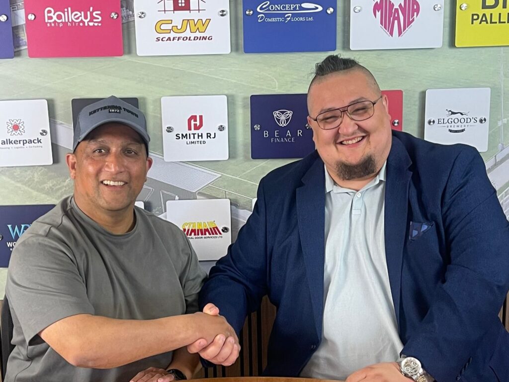 Two men shake hands and smile at the camera; left man in gray t-shirt and cap, right man in a navy blazer, with sponsor logos on the wall behind them.