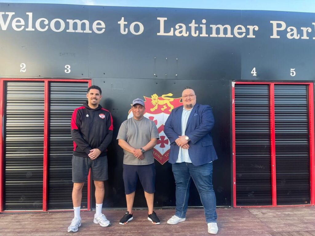 Three men posing in front of a black wall that reads 'Welcome to Latimer Park' with a crest between them, flanked by red-and-black storage bays numbered 2–5.