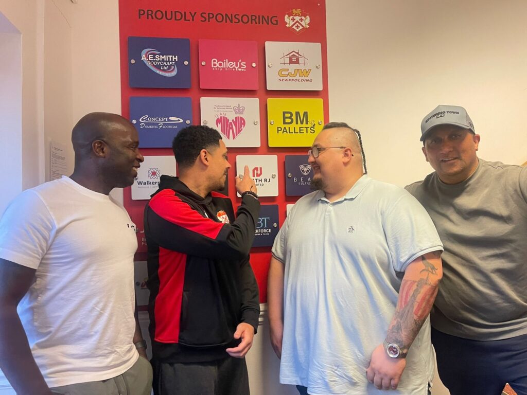 Four men stand together in front of a wall displaying sponsor logos; the man in black and red gestures as he talks to the man in a light blue polo.