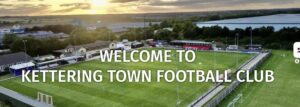 Aerial view of Kettering Town Football Club stadium at sunset with a large 'Welcome to Kettering Town Football Club' banner across the field.