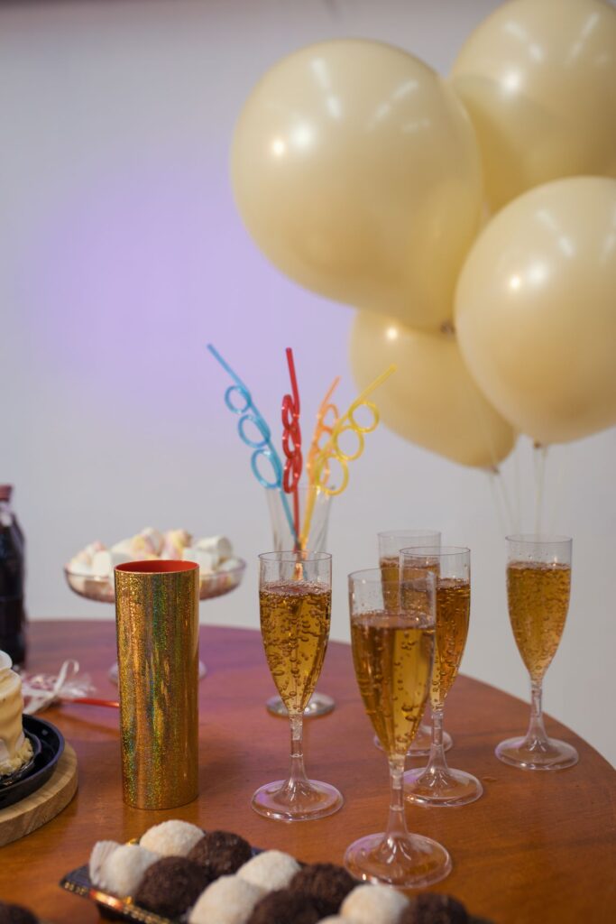 Celebration table with champagne flutes, gold glitter cup, colorful swizzle sticks, and beige balloons in the background.