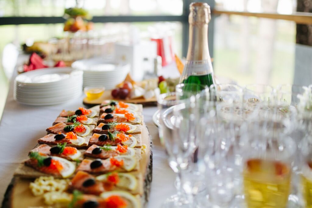 Buffet table with assorted canapés topped with salmon, lemon, olives and herbs, a champagne bottle in the background, and a stack of plates with empty glasses nearby.