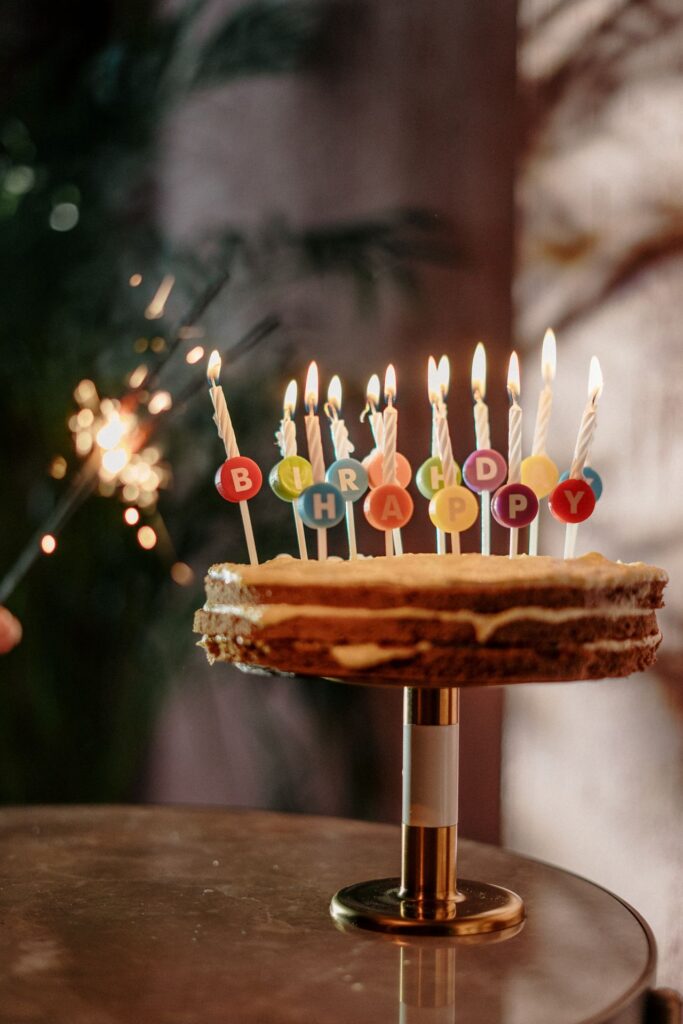 Birthday cake on a pedestal with lit striped candles and colorful round toppers spelling BIRTHDAY