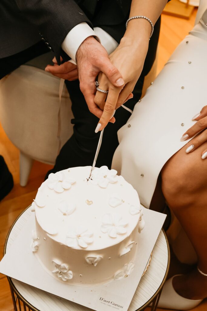 Couple's hands cut a white wedding cake decorated with small flowers, both wearing rings (wedding bands).