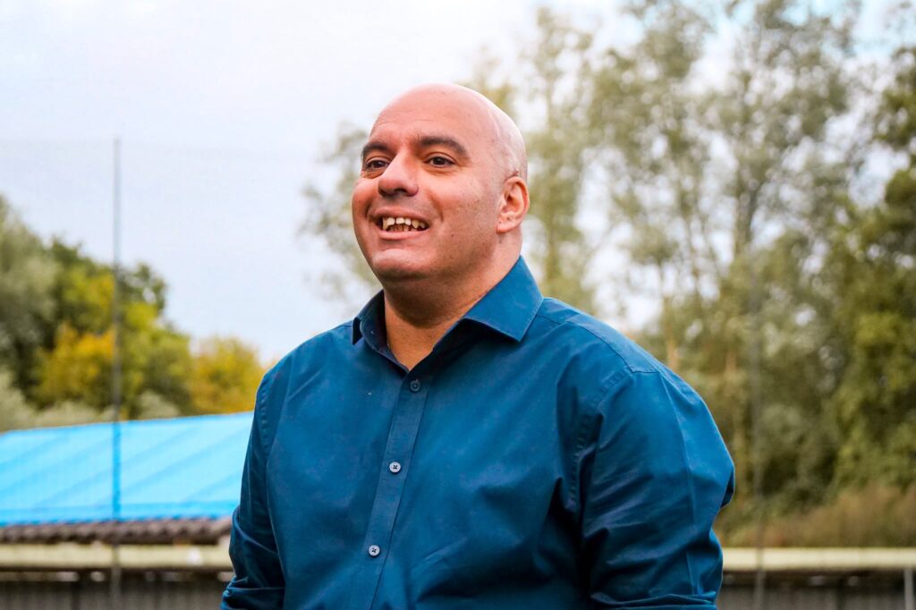 KTFC_Dave_Hennessey | Kettering Town FC Smiling bald man in a blue button-down shirt outdoors, with a blue fence and trees in the background.