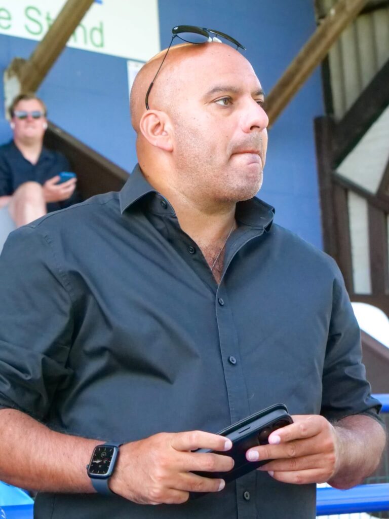 Man in a dark button-down shirt holding a tablet, wearing sunglasses on his head, outdoors with another person in the background.