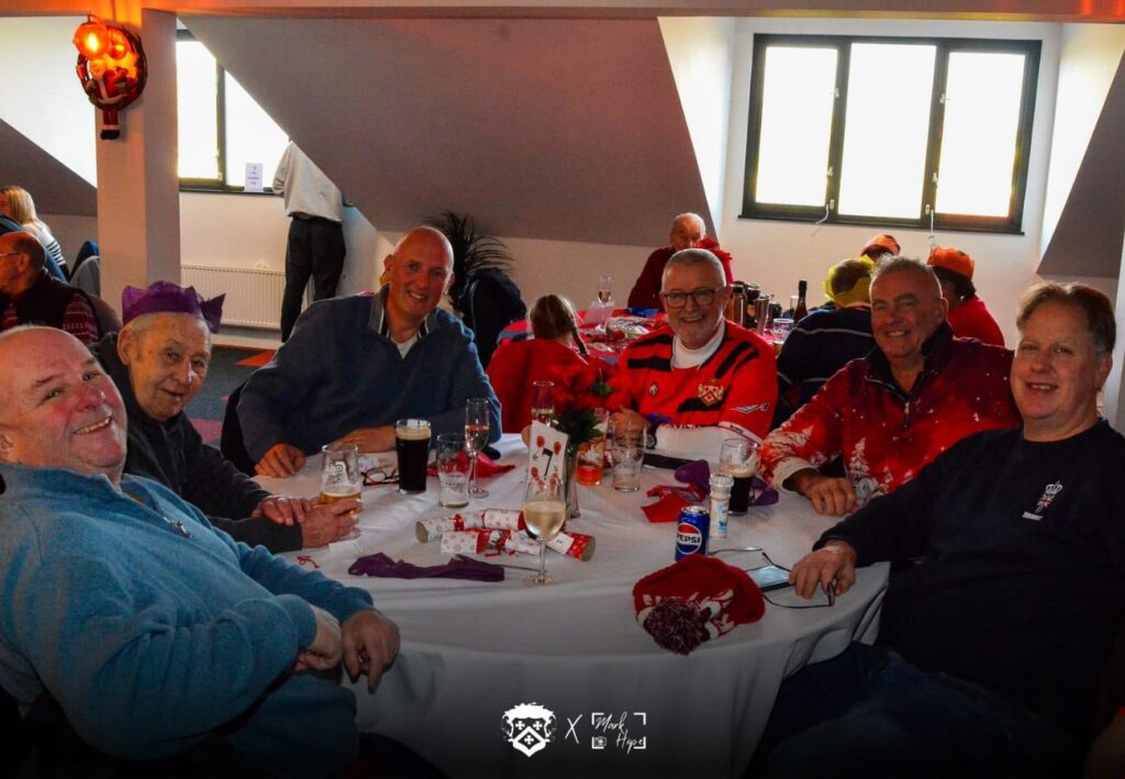 Group of men seated around a round table, smiling, with drinks and festive decorations at a holiday party.