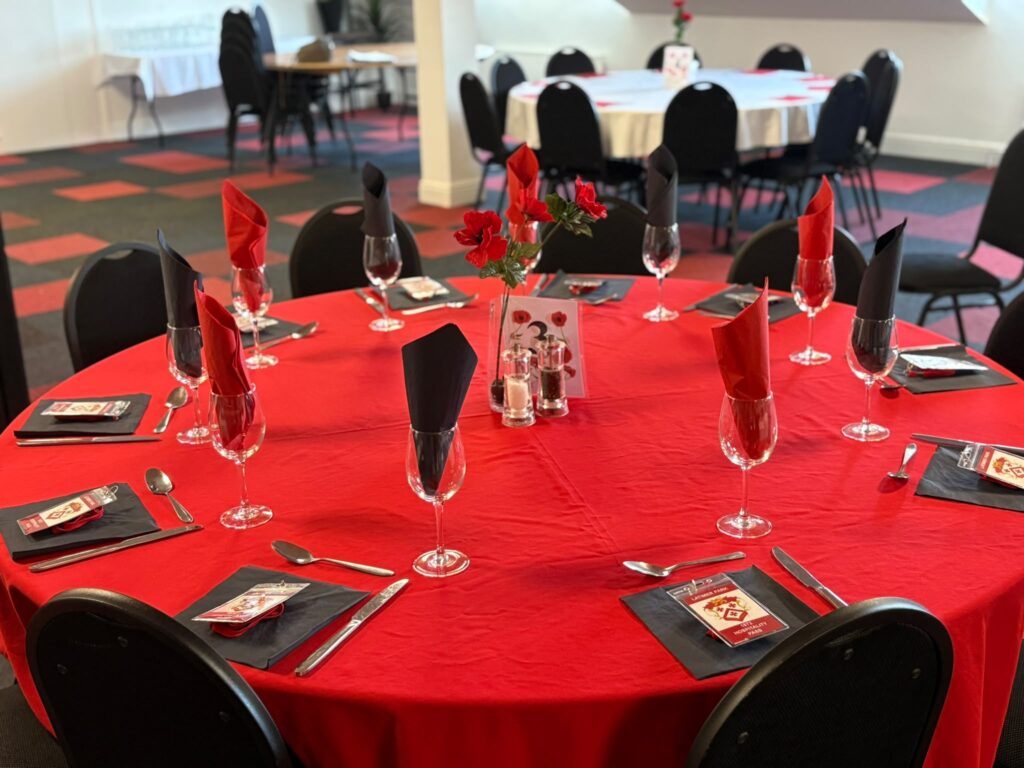Round banquet table with red tablecloth, black napkins, and wine glasses; center vase with red flowers in a decorated event space.