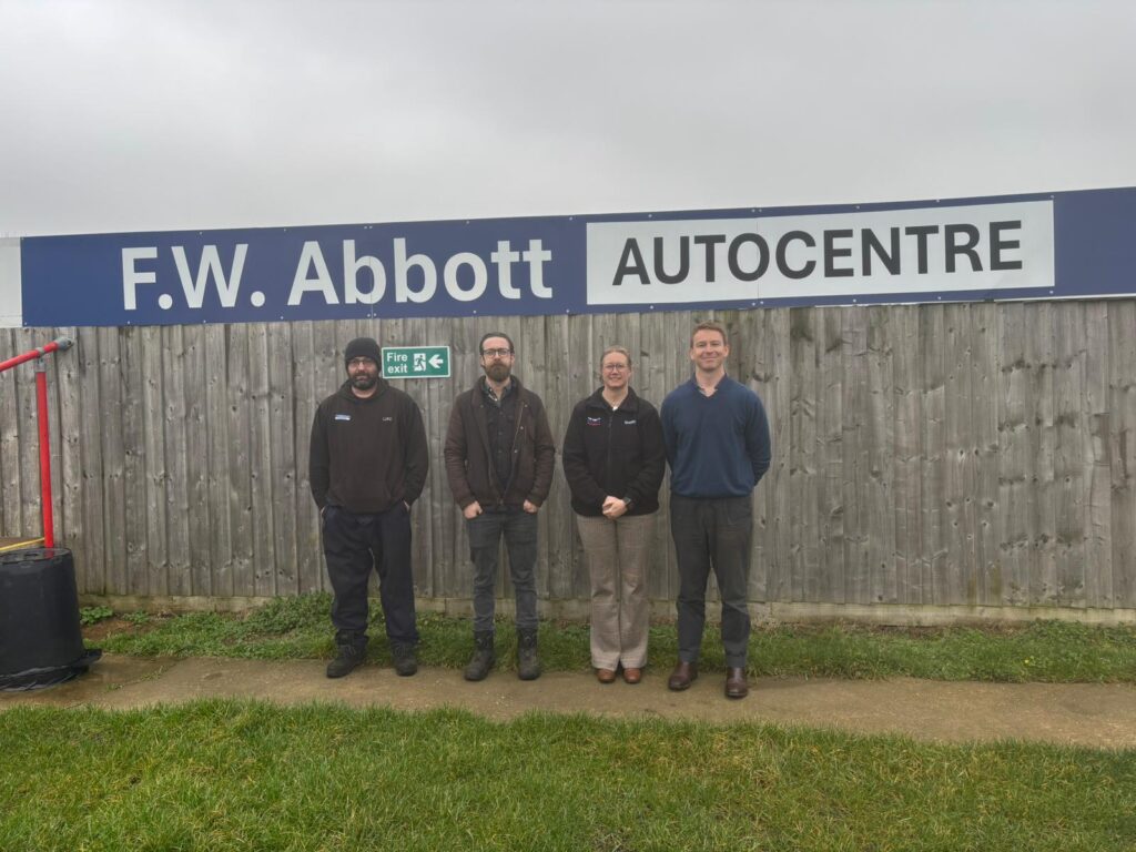 KTFC_FWABOTT2 | Kettering Town FC Four people stand side by side in front of a wooden fence and a sign that reads'F.W. Abbott Autocentre'—outdoor group photo.