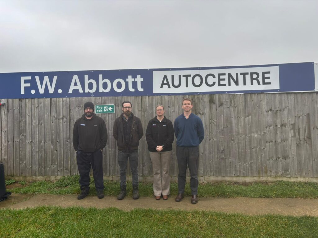 Four people stand side by side in front of a wooden fence beneath a large blue sign that reads 'F.W. Abbott AUTOCENTRE' on a cloudy day