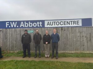 Four people stand side by side in front of a wooden fence beneath a large blue sign that reads 'F.W. Abbott AUTOCENTRE' on a cloudy day
