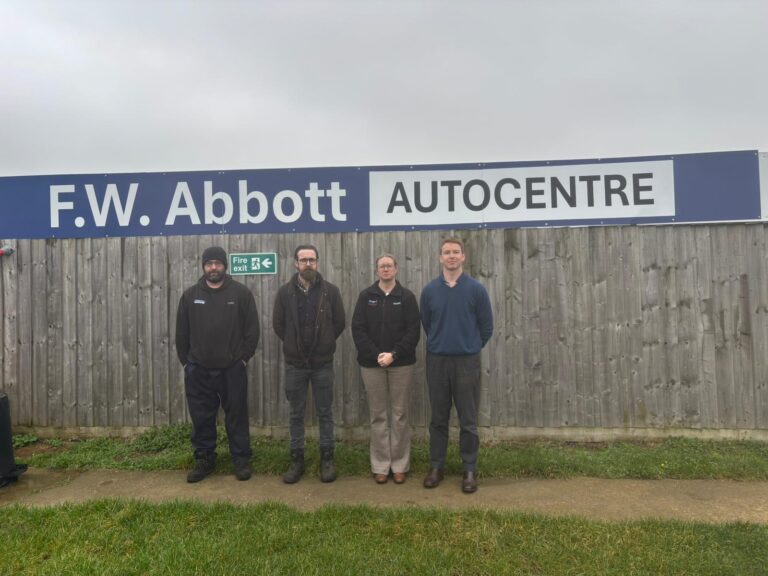 KTFC_FWABOTT4 | Kettering Town FC | Kettering Town FC Four people stand side by side in front of a wooden fence beneath a large blue sign that reads 'F.W. Abbott AUTOCENTRE' on a cloudy day