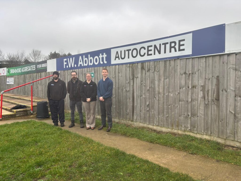KTFC_FWABOTT7 | Kettering Town FC Group of four adults standing in front of a wooden fence with a large'F.W. Abbott AUTOCENTRE' sign behind them, outdoors on a cloudy day.