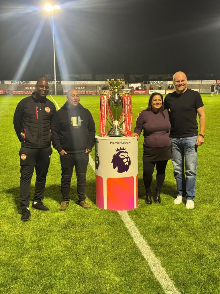 KTFC_Floodlights | Kettering Town FC | Kettering Town FC Four people pose with a Premier League trophy on a football pitch at night under bright floodlights, smiling.