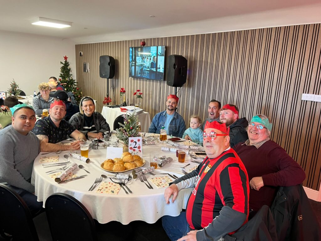 Group of people around a holiday buffet table, some wearing paper crowns, Christmas tree and decorations in the background.